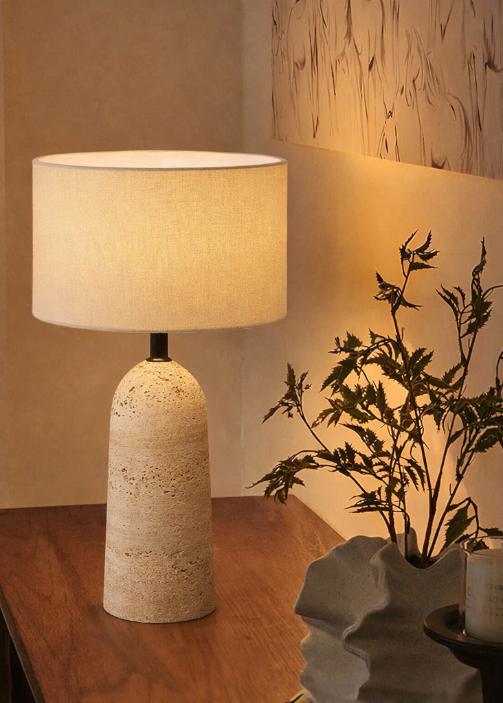 Table lamp with textured base and beige shade on a wooden surface, next to a plant and candle.