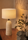 Table lamp with textured base and beige shade on a wooden surface, next to a plant and candle.